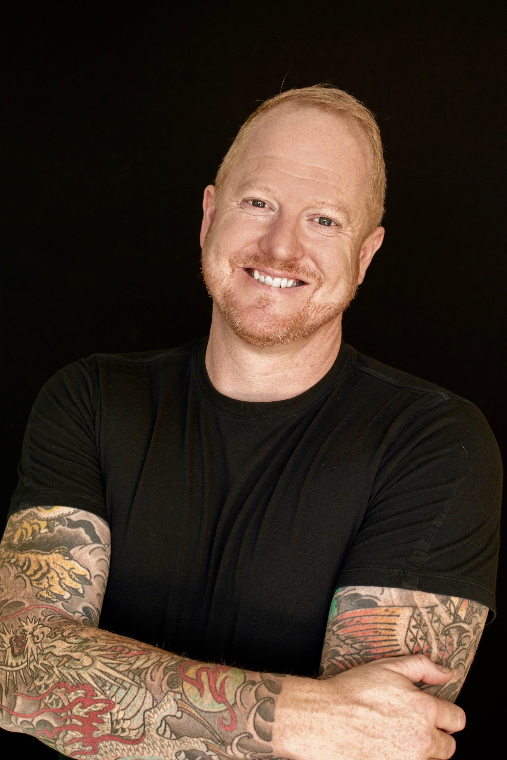 Nikolas Badminton, a white man with short red hair, beard, and moustache, is standing in front of a black background with his tattooed arms crossed, wearing a black t-shirt.