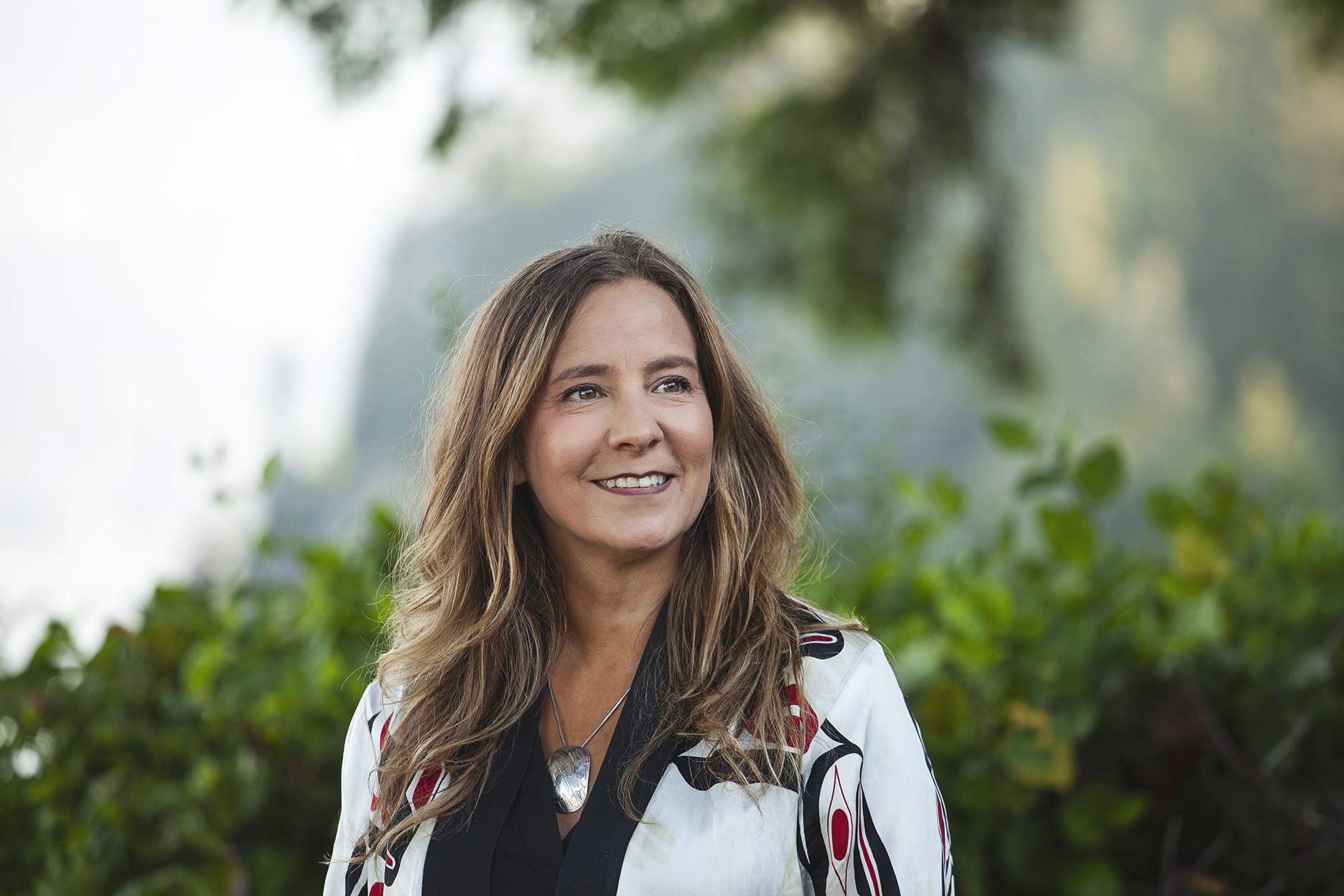 Teara Fraser stands in front of greenery, looking to the left of the camera. Teara is a Métis woman of Cree ancestry with long wavy brown hair. She is wearing a white, red, and black jacket over a black shirt and silver necklace with a silver pendant.