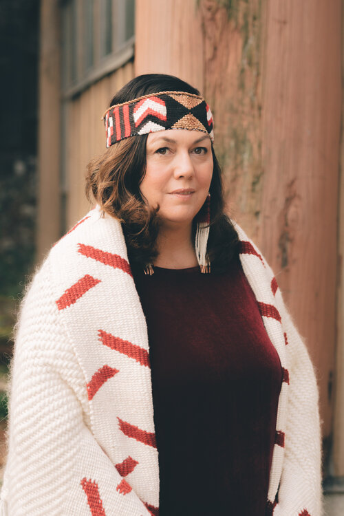 Ta7talíya Michelle Nahanee, stands in front of a wooden building, looking to the left of the camera. Ta7talíya Michelle is a First Nations woman with curly brown hair under a beaded headband. She wears long beaded earrings, a deep red velvet shirt, and a cream blanket with red accents.